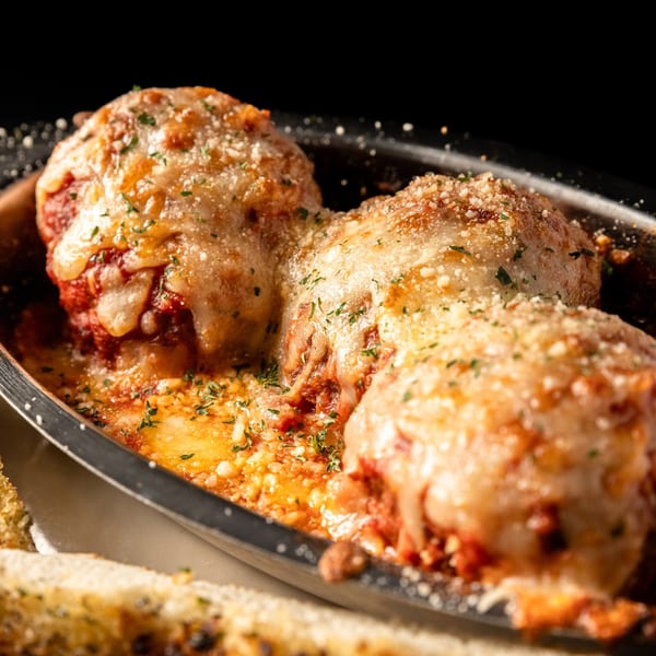 Three large, cheesy meatballs in red sauce, garnished with herbs, served in a dark metal dish.