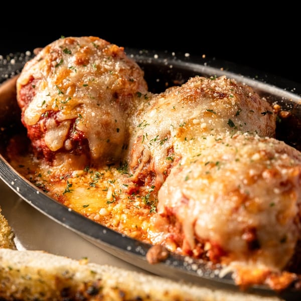 Three meatballs covered in melted cheese and red sauce, garnished with parsley, in a metal dish.
