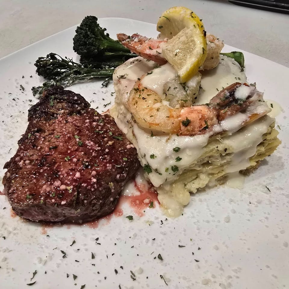 Seared steak, creamy lasagna, and broccolini, garnished with shrimp, lemon, and herbs on a white plate.