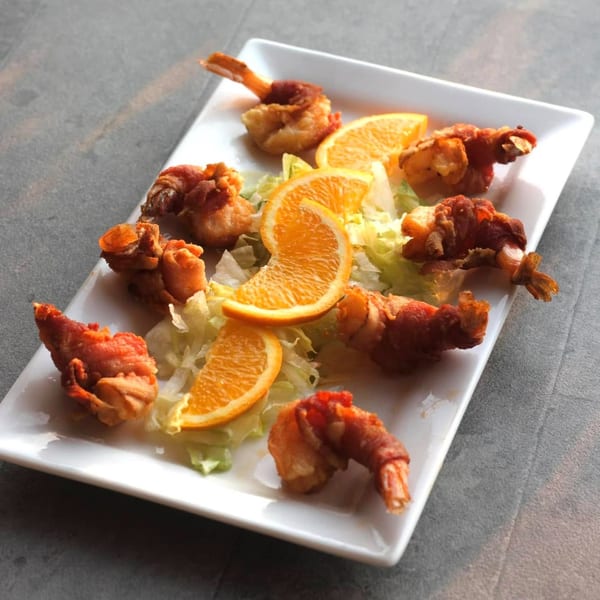 A white rectangular plate holds several fried shrimp wrapped in bacon, garnished with lettuce and orange slices.