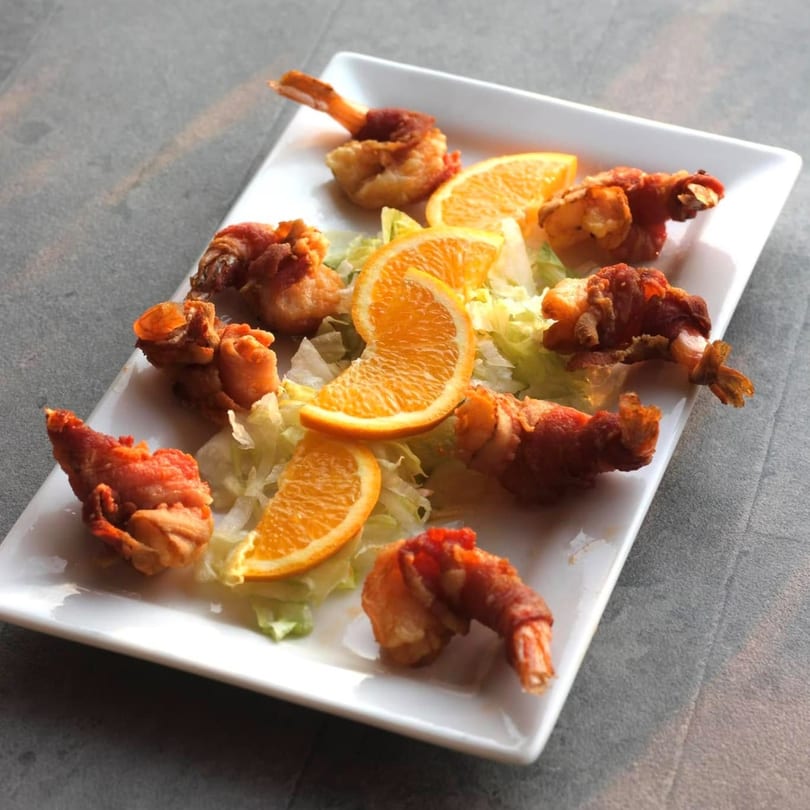 A white rectangular plate holds several fried shrimp wrapped in bacon, garnished with lettuce and orange slices.