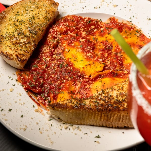 Plate of Gusto Dolce's ravioli with red sauce, parmesan, parsley, and garlic bread, with a drink.