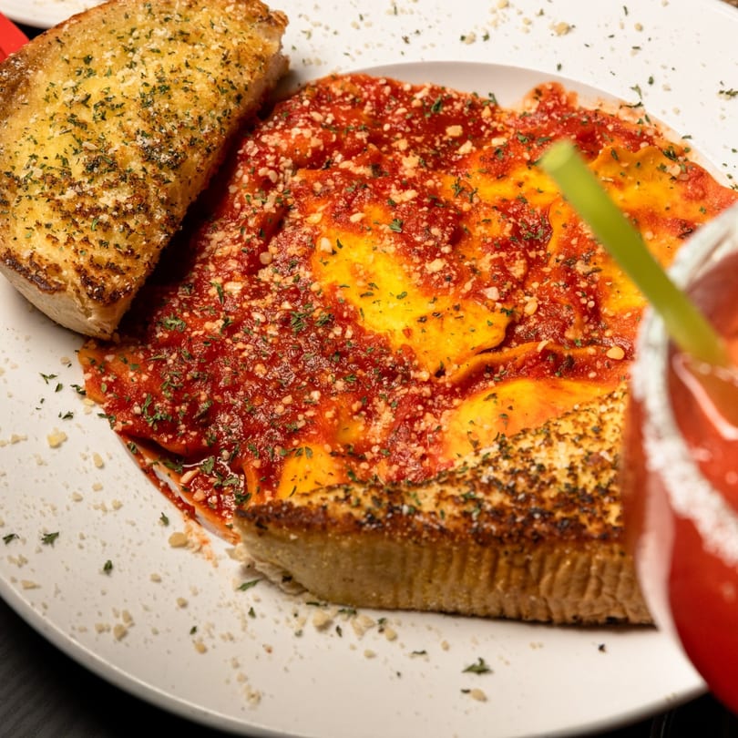 Plate of Gusto Dolce's ravioli with red sauce, parmesan, parsley, and garlic bread, with a drink.