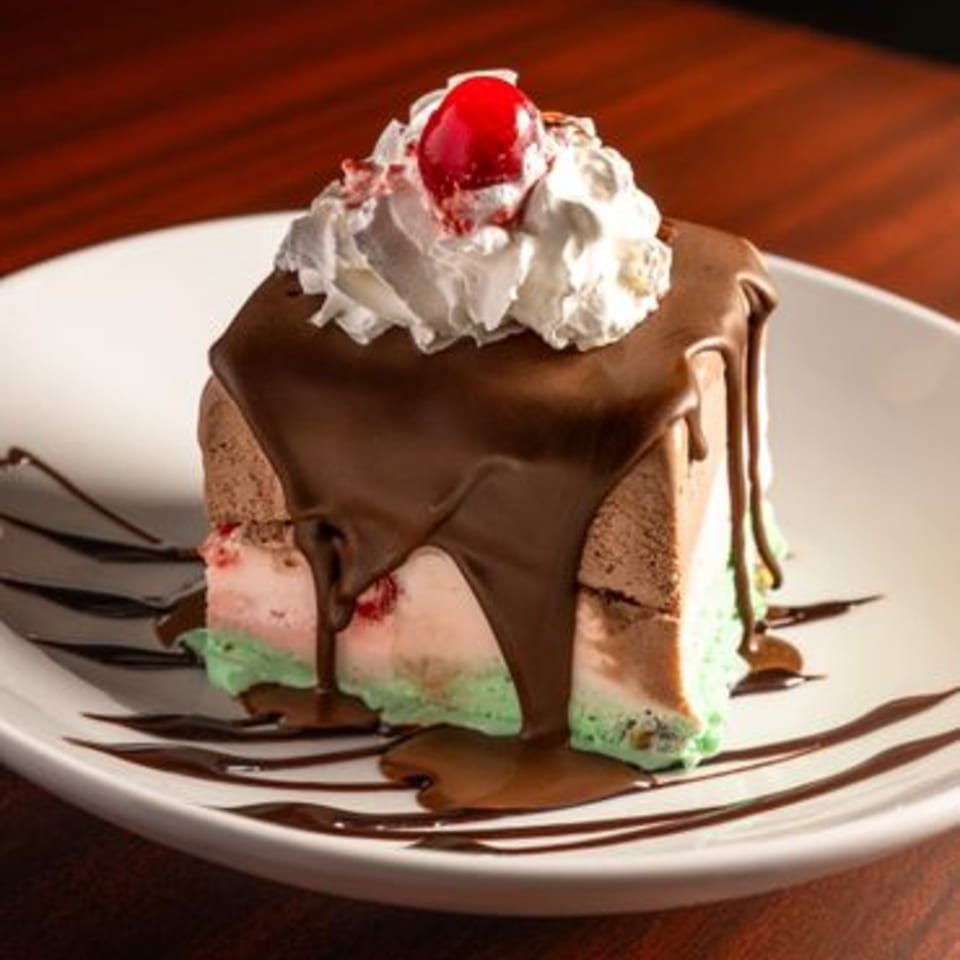 A slice of spumoni ice cream cake with chocolate sauce, whipped cream, and a cherry on top.