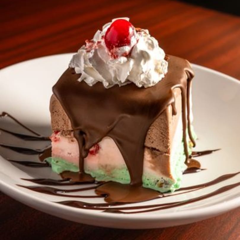A slice of spumoni ice cream cake with chocolate sauce, whipped cream, and a cherry on top.