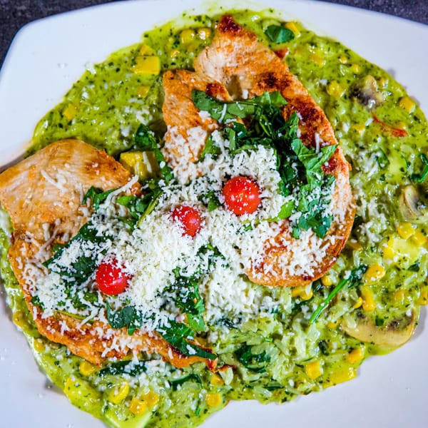 Grilled chicken breasts over green risotto with corn, mushrooms, cherry tomatoes, and grated cheese.
