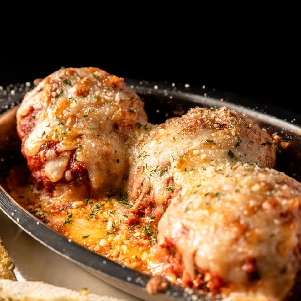Three large meatballs covered in melted cheese, tomato sauce, and parsley in a dark dish.