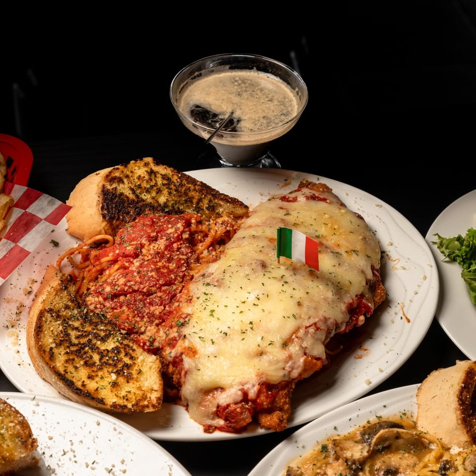 Chicken parmesan with spaghetti, garlic bread, and an Italian flag pick, alongside a dark drink.
