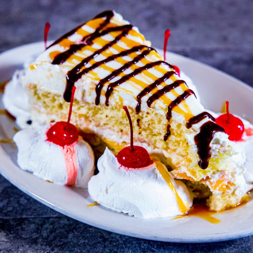 Slice of Italian cake with white frosting, chocolate, caramel drizzle, whipped cream, and cherries.